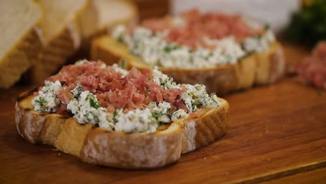 Tasty-homemade-bruschetta-with-ham-on-white-cheese-served-in-a-wooden-cutting-board---macro,-close-up-slider-movement