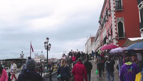 Gente-Y-Turistas-Caminando-Hacia-Un-Puente-Que-Cruza-El-Canal-De-Venecia-Al-Lado-De-La-Tienda-De-La-Calle-En-Un-Día-Lluvioso-Nublado