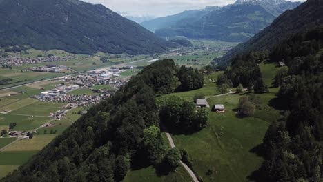 Drone-tilts-up-while-flying-over-a-small-green-hill-with-two-sheds-next-to-a-big-forest-and-mountains-in-the-Swiss-alps-near-the-lake-of-Walen
