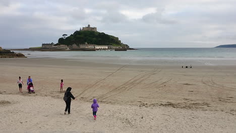 Mothers-and-their-children-walking-on-the-beach-of-Marazion-in-front-of-the-english-medioeval-castle-and-church-of-St-Michael's-Mount-in-Cornwall-on-a-cloudy-spring-day,-4k-footage