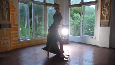 A-mysterious-young-woman-with-shaved-head-is-dancing-sensually,-spinning-her-arms,-legs-and-dress-dramatically-while-being-lit-by-a-harsh-light-behind-her