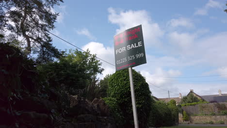 Low-Angle-Static-Shot-of-British-‘For-Sale’-Real-Estate-Sign-on-Sunny-Summer’s-Day-with-Foliage-Blowing-in-the-Breeze-in-the-Background-in-Slow-Motion