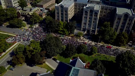 Large-crowd-marches-by-stone-buildings-in-Toronto,-aerial-drone-pan
