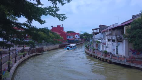 Tourist-Boat-cruising-into-a-narrow-Melaka-river-with-people-in-the-boat,-houses-and-tress-on-both-side-of-the-river
