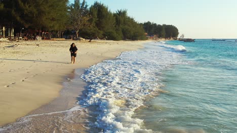 Girl-walking-alongside-white-waves-of-blue-sea-on-white-sand-of-exotic-beach-of-tropical-island-in-Bali