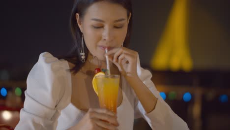 Stunning-Asian-woman-with-an-orange-cocktail-at-a-rooftop-bar-and-the-city,-bokeh-lights-in-the-background