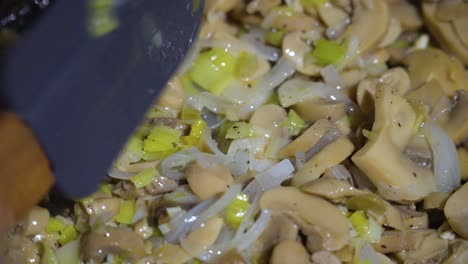 Close-up-of-chopped-mushrooms,-leek-and-onion-in-frying-pan-stirred-with-spatula