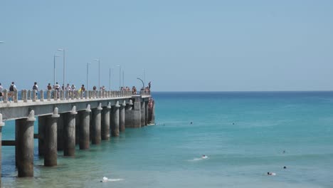 Many-people-walking-on-Porto-Santo-city-island-pier-with-tourists-swimming-in-turquoise-ocean-sea-water-on-hot-summer-blue-sunny-sky-cloudless-day,-Portugal,-static