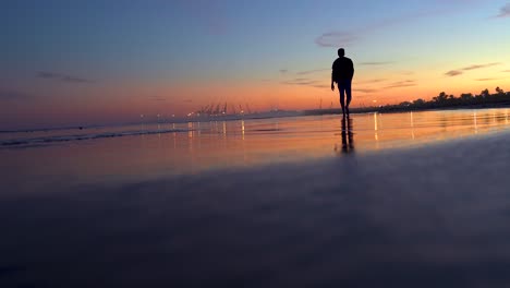 Man-walks-towards-the-horizon-on-beach-at-sunset,-backlit-with-an-orange-sky-and-blue-the-beach-inclined-with-the-sand-and-the-reflection-of-water-on-the-slope