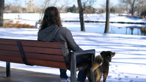 Man-with-dark-long-hair-sitting-at-a-bench-petting-his-dog-in-Winter-at-a-park