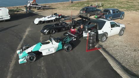 Man-Sitting-Close-To-The-Racing-Car-Parked-On-The-Road-Side-At-The-Hill-In-Imtahleb-Malta-With-Pile-Of-Wheels-For-Reserve---Aerial-Shot