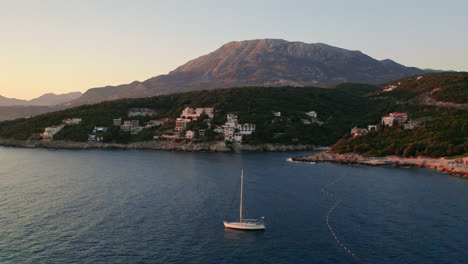 Static-aerial-shot-of-a-yacht-anchoring-in-a-bay-in-Montenegro-in-the-evening-with-a-small-coastal-city-and-a-large-mountain-in-the-background-and-a-clear-sky-above