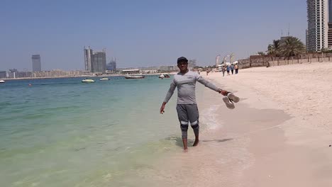 Adult-Asian-male-walks-on-the-beach-and-turns-around,-hands-raised-wide-open-happily-and-walks-back-away-again-in-Jumeirah-beach-sunny-day