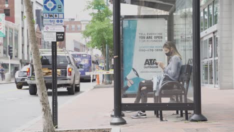 Woman-sits-on-bench-in-glass-bus-station-looking-at-phone-and-wearing-face-mask