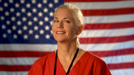 Medium-tight-portrait-of-nurse-looking-to-the-left-and-then-at-camera,-relieved,-happy-and-nodding-her-head-with-American-flag-behind-her