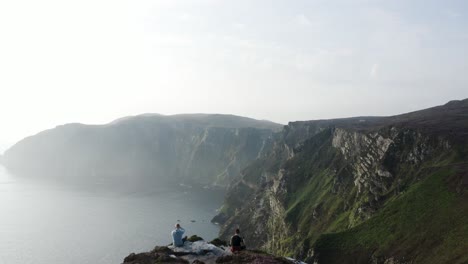 Zwei-Personen-Sitzen-Auf-Der-Klippe-Und-Blicken-Auf-Die-Bergkette-Am-Horn-Head-In-Donegal,-Irland