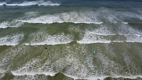 Birds-eye-drone-shot-of-Muizenberg-beach,-Cape-Town---drone-is-descending-over-surfers-trying-to-catch-a-wave