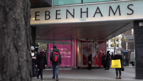 A-slow-motion-left-to-right-camera-truck-technique-passes-a-tree-trunk-and-reveals-people-entering-and-exiting-Debenhams-department-store-on-Oxford-Street-as-others-walk-by-carrying-shopping-bags