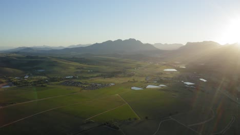 Sun-rays-sunrise-lens-flare-over-vineyard-landscape,-mountains-aerial,-Stellenbosch,-Longlands