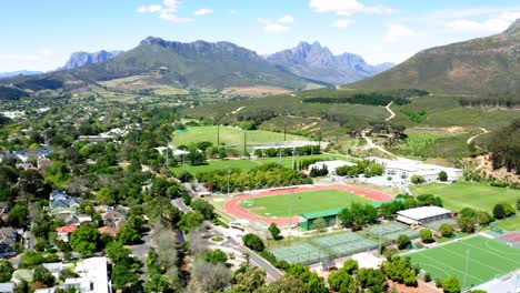 Aerial-drone-rotating,-university-college-sports-athletics-stadium,-track-and-field,-tennis-courts,-mountains,-trees-and-neighbourhood-in-background,-Stellenbosch,-Coetzenburg