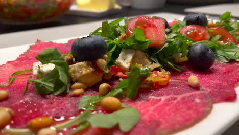Close-up-tilt-down-shot-of-beautiful-served-carpaccio-meat-with-arugula,-tomatoes,-blueberries-and-roasted-seeds