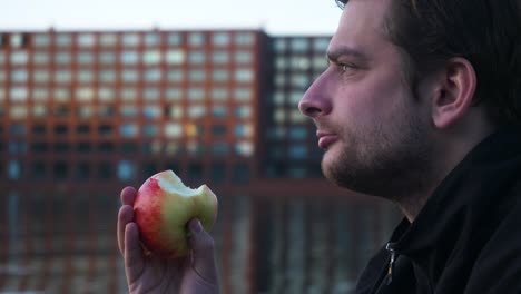 Close-up-of-a-young-man-eating-a-red-apple-and-looking-into-the-distance-on-a-pier-during-the-evening