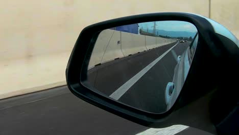 View-in-rear-mirror-from-car-on-highway,-personal-transportation,-car-driving-on-motorway-along-concrete-wall