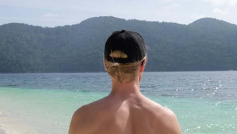 Ultra-slow-motion-shot-behind-young-caucasian-man-with-cap-walking-on-stunning-beach-in-Thailand