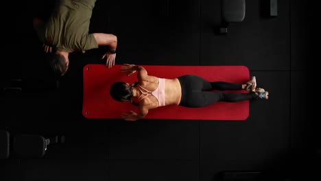Top-down-drone-shot-of-sporty-woman-doing-push-ups-in-gym-with-personal-trainer