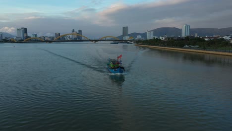 Aerial-orbit-shot-around-a-fishing-boat-at-sunset-with-the-city-skyline-of-Vietnam-in-background