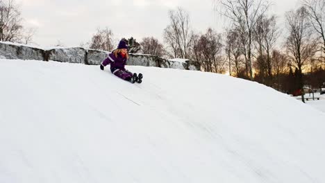 Girl-playing-outside,-sliding-down-a-snowy-ice-hill-on-her-bottom-in-crispy-winter-weather
