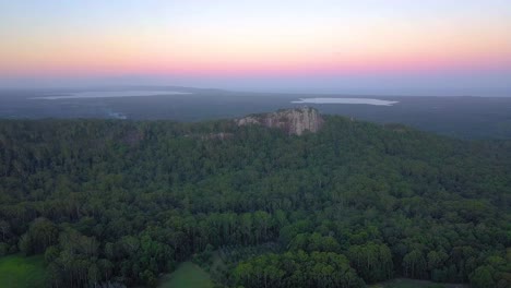 Rising-aerial-view-of-Mount-Tinbeerwah-at-sunset-with-tropical-forest,-ocean-and-pink-sky