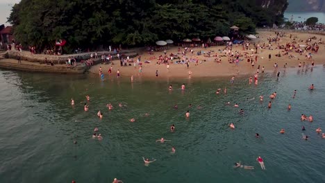 Aerial-shot,-circling-left-over-swimmers-at-beach-excursion-on-Ha-Long-Bay-cruise-late-afternoon,-tilt-up-at-end-to-reveal-tall-island-cliff
