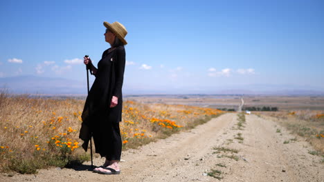 An-old-woman-in-a-dramatic-black-dress-and-sun-hat-on-a-dirt-road-under-blue-skies-with-flowers-and-wind-blowing-in-slow-motion