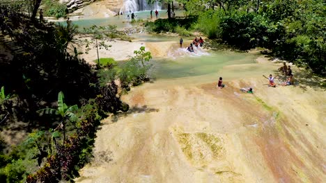 A-beautiful-waterfall-cascades-down-a-cliff-into-a-beautiful-turquoise-pool-below-in-the-middle-of-a-lush-green-jungle-in-Bohol,-Philippines-while-locals-enjoy-the-cool-relaxing-water