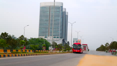 A-red-metro-bus-coming-on-the-road,-crossing-the-camera,-low-angle,-close-up,-Behind-a-tri-building-tower-and-couple-of-buildings,-beautiful-street-lights-and-greenery-on-the-road