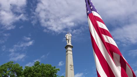 Civil-war-monument-statue-on-memorial-day-in-a-cemetery-under-a-cloudy-blue-sunny-sky-and-the-American-flag-waving-on-a-windy-day