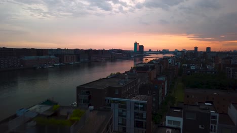 Flying-over-Amsterdam-skyline-by-sunset-by-drone-over-the-IJ-river-near-central-station-with-beautiful-red-sky-and-white-train-heading-to-train-station