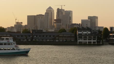 Londons-Finanzviertel-Canary-Wharf-Mit-Einem-Ausflugsboot,-Das-In-Der-Abenddämmerung-Auf-Der-Themse-Vorbeifährt