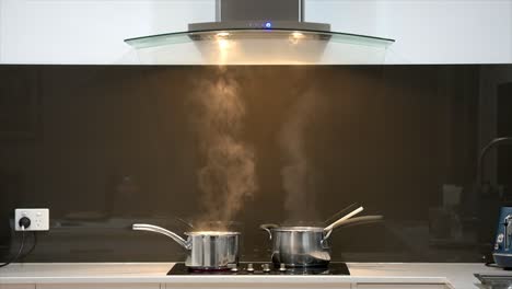 Wide-shot-of-a-pair-of-simmering-pans-on-a-cook-top-with-steam-rising-into-a-rangehood