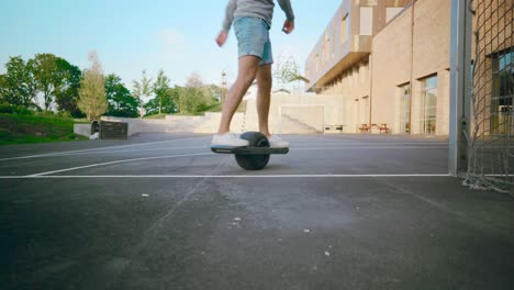 Young-mand-boarding-One-Wheel-skating-away-in-schoolyard-with-threes-and-blue-sky-during-summer-shot-from-frogs-eye-perspective