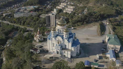 Slow-descent-pivot-aerial-shot-of-an-orthodox-church-with-blue-roof-and-golden-domes,-located-on-top-of-the-hill-with-port-and-city-building-in-the-background,-on-a-bright,-clear,-sunny-day