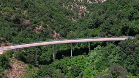 Eine-Geschwungene-Balkenbrücke-über-Einen-Steilen,-Mit-Bäumen-Und-Büschen-Bedeckten-Bergrücken-In-Der-Nähe-Von-Milocher,-Montenegro,-Mit-Einem-Weißen-Auto-Und-Einem-Weißen-Lieferwagen,-Die-An-Einem-Sonnigen-Sommertag-Vorbeifahren,-Luftaufnahme