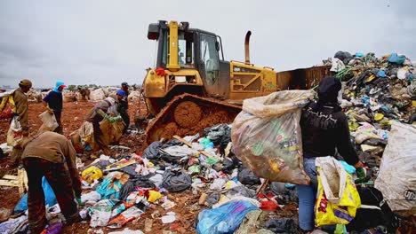 big-pile-of-trash-moved-by-a-bulldozer,-while-unidentified-people-pick-up-something-usable,-on-a-landfill-site