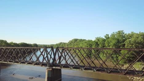 Old-truss-railroad-bridge-above-narrow-river-in-rural-landscape-with-flock-of-black-birds-flying-in-foreground,-aerial-overhead-approach