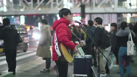 A-Male-Guitarist-Busking-On-The-Street-Outside-Shinjuku-Station-In-Tokyo,-Japan-At-Night---medium-slowmo-shot