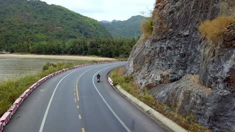 Woman-on-a-moped-riding-on-an-island-road-near-the-coastline,-Aerial-follow-behind-shot