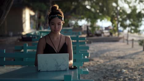 Young-influencer-girl-working-on-laptop-at-beach-lit-by-sunset,-Bali