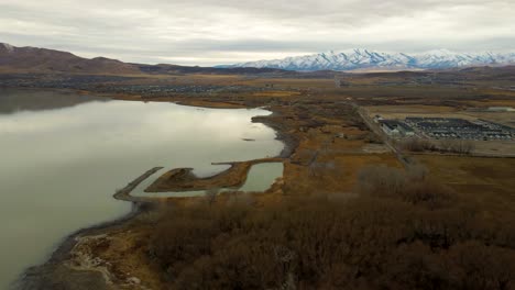 Flying-high-over-a-lake-and-shoreline-with-mountains-in-the-distance-on-an-overcast,-winter-day
