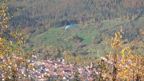 Parapente-Sobrevolando-Una-Pequeña-Ciudad-Y-Un-Impresionante-Paisaje-Otoñal-En-El-Teufelsmühle-En-La-Selva-Negra,-Alemania